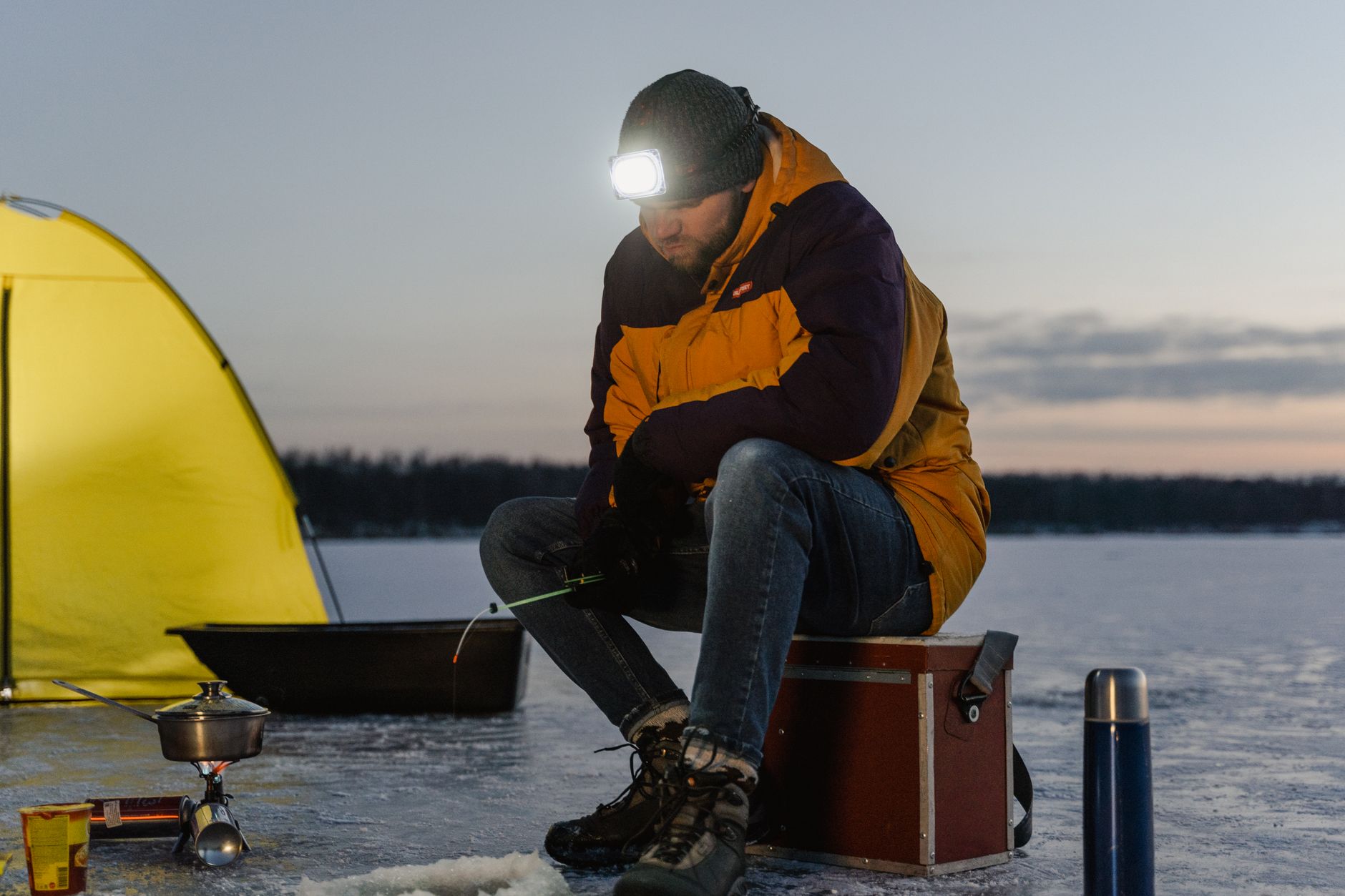 A man ice fishing on a frozen lake, surrounded by camping gear at dusk.