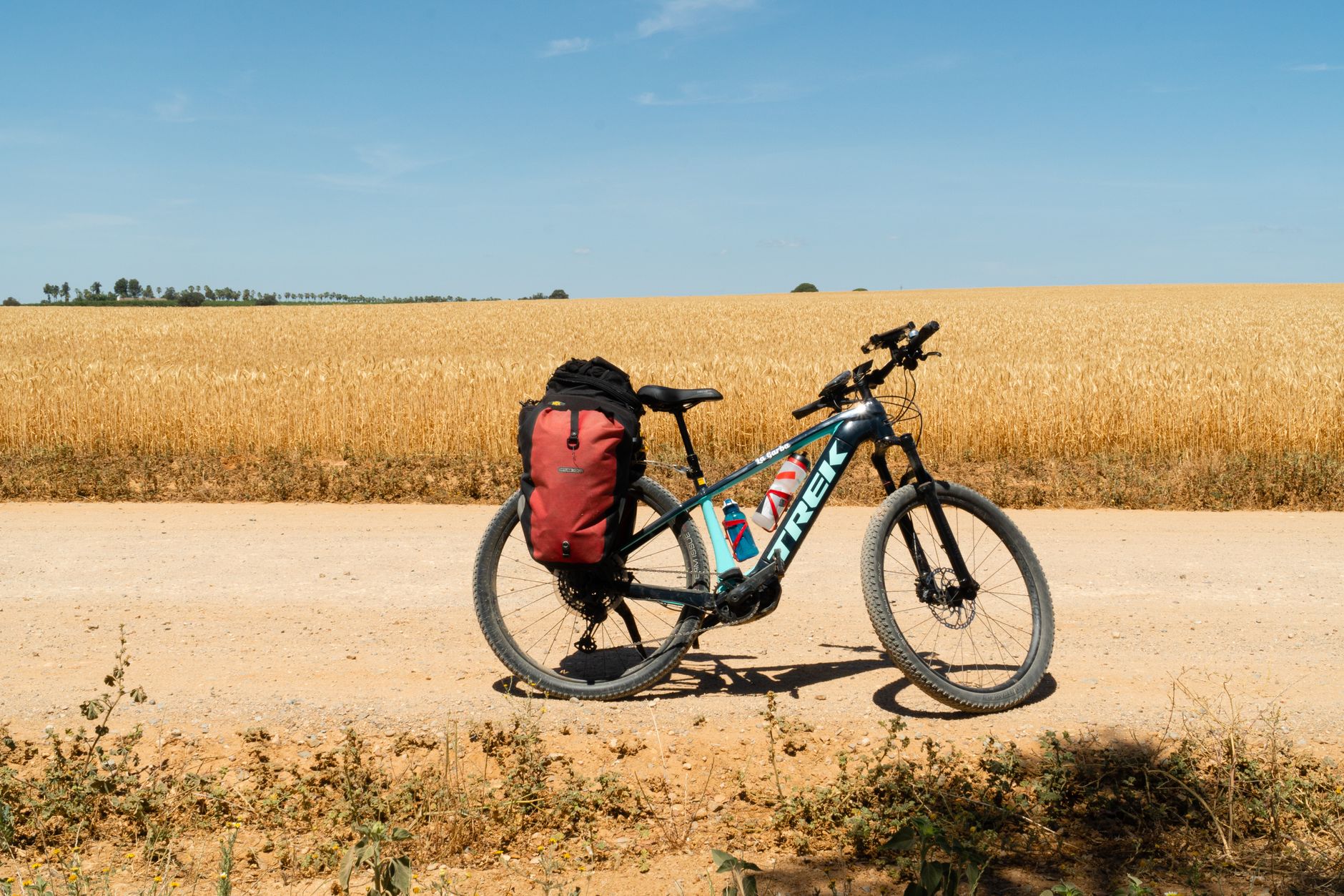 A mountain bike with a red backpack parked on a rural dirt road next to expansive golden fields under a clear blue sky.