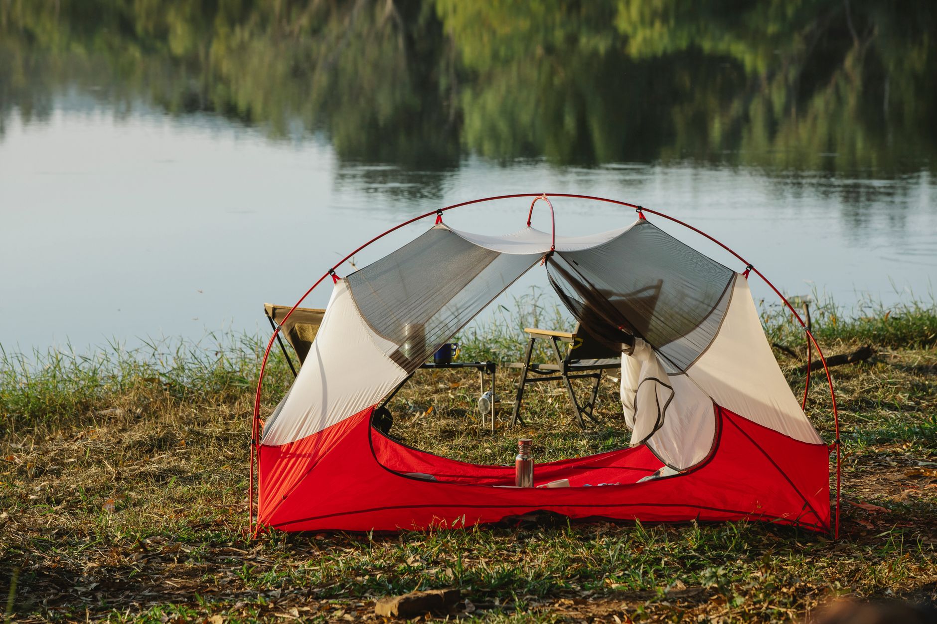 Camping tent and chairs placed on grassy shore of peaceful lake surrounded by lush green forest during romantic picnic on sunny day