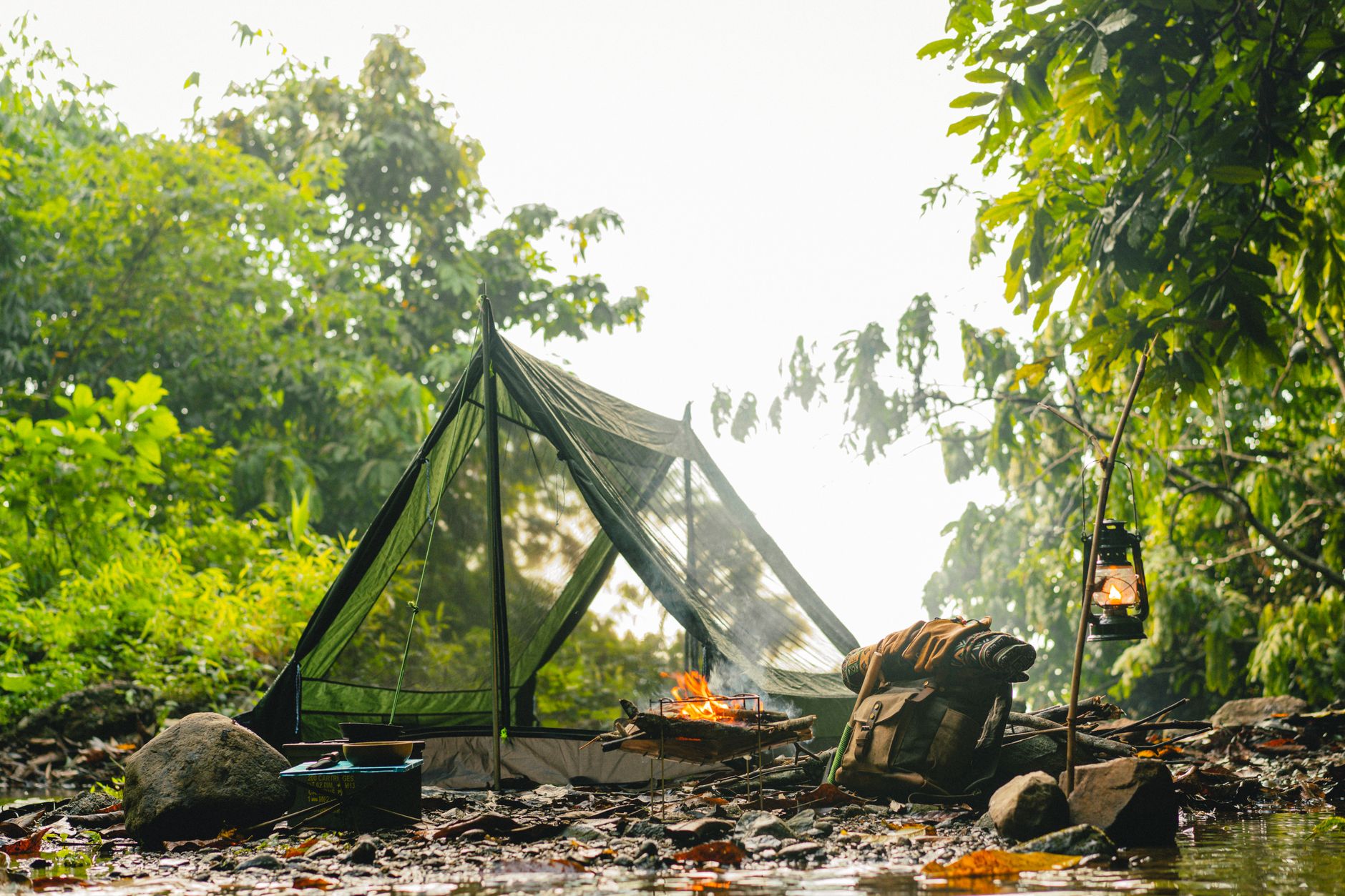 A serene camping site with a tent, campfire, and kerosene lamp in lush greenery during daylight.