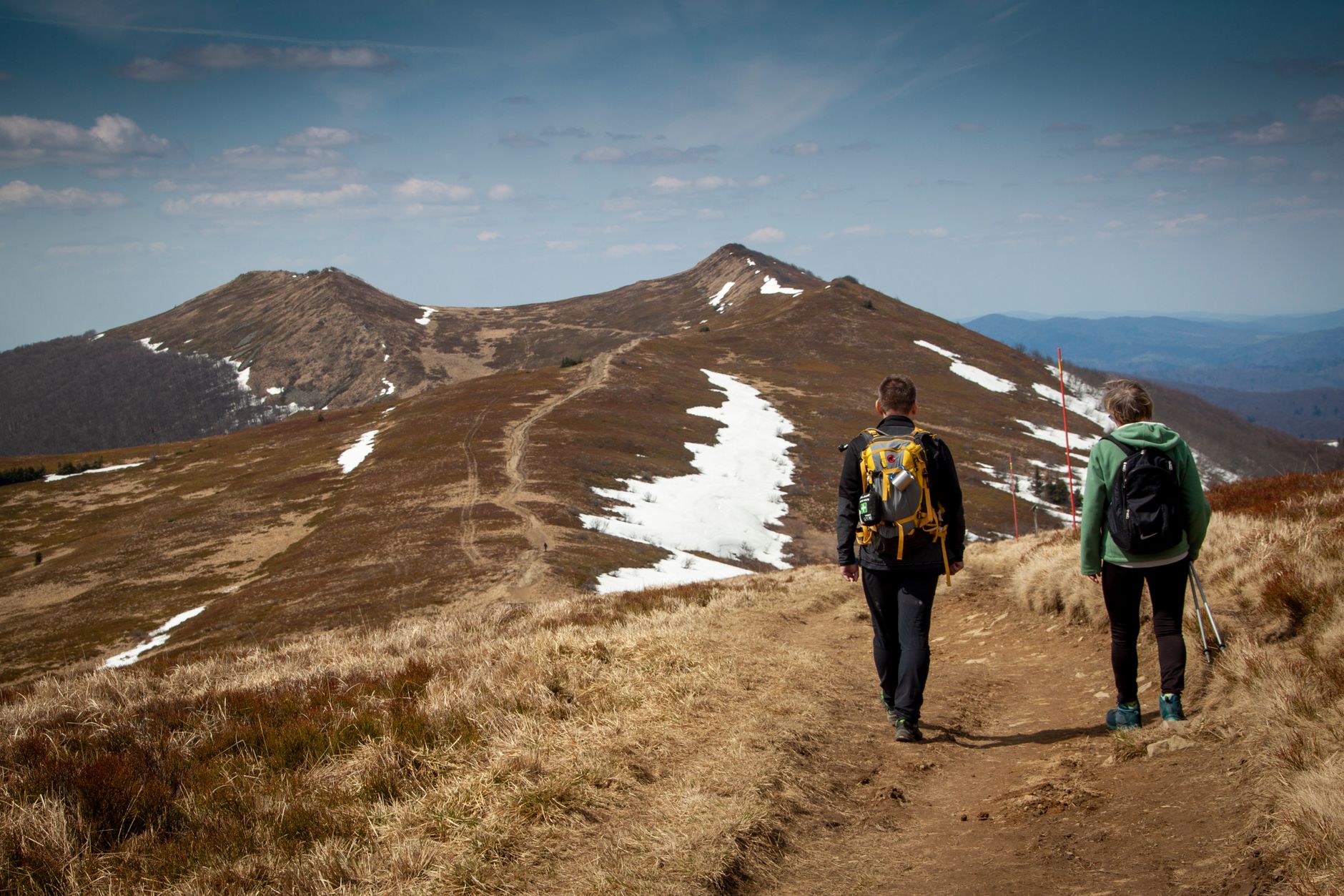 Two hikers traverse a scenic mountain trail with patches of snow in early spring.
