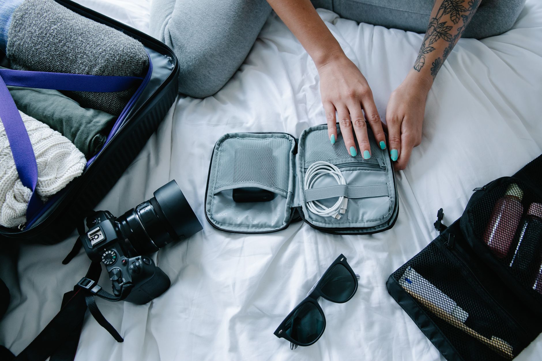 Woman with tattoos packs electronics, camera, and sunglasses in a suitcase on a bed.