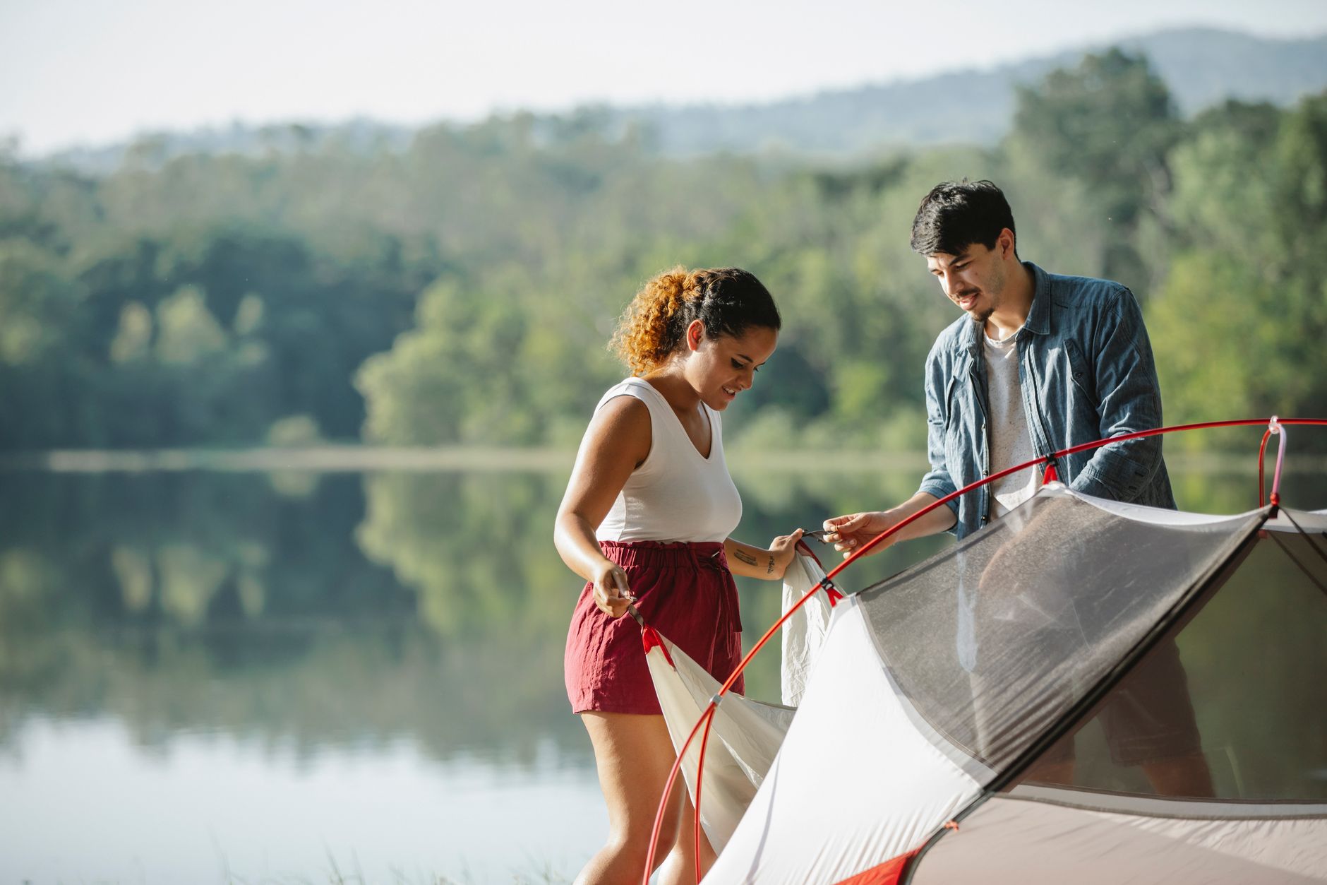 Multiethnic travelers putting up tent against trees reflecting in lake during summer trip in sunlight