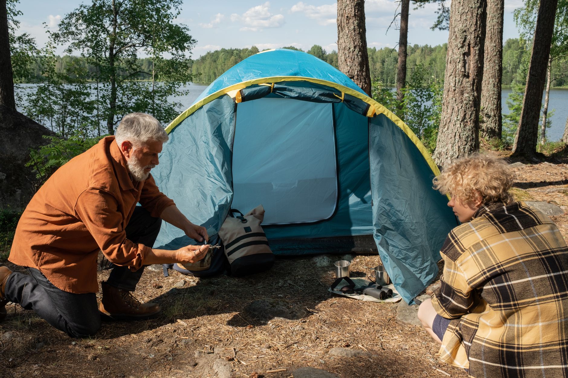 An elderly man and a teenager pitch a tent by the lake in a serene forest setting, bonding during a summer camping trip.