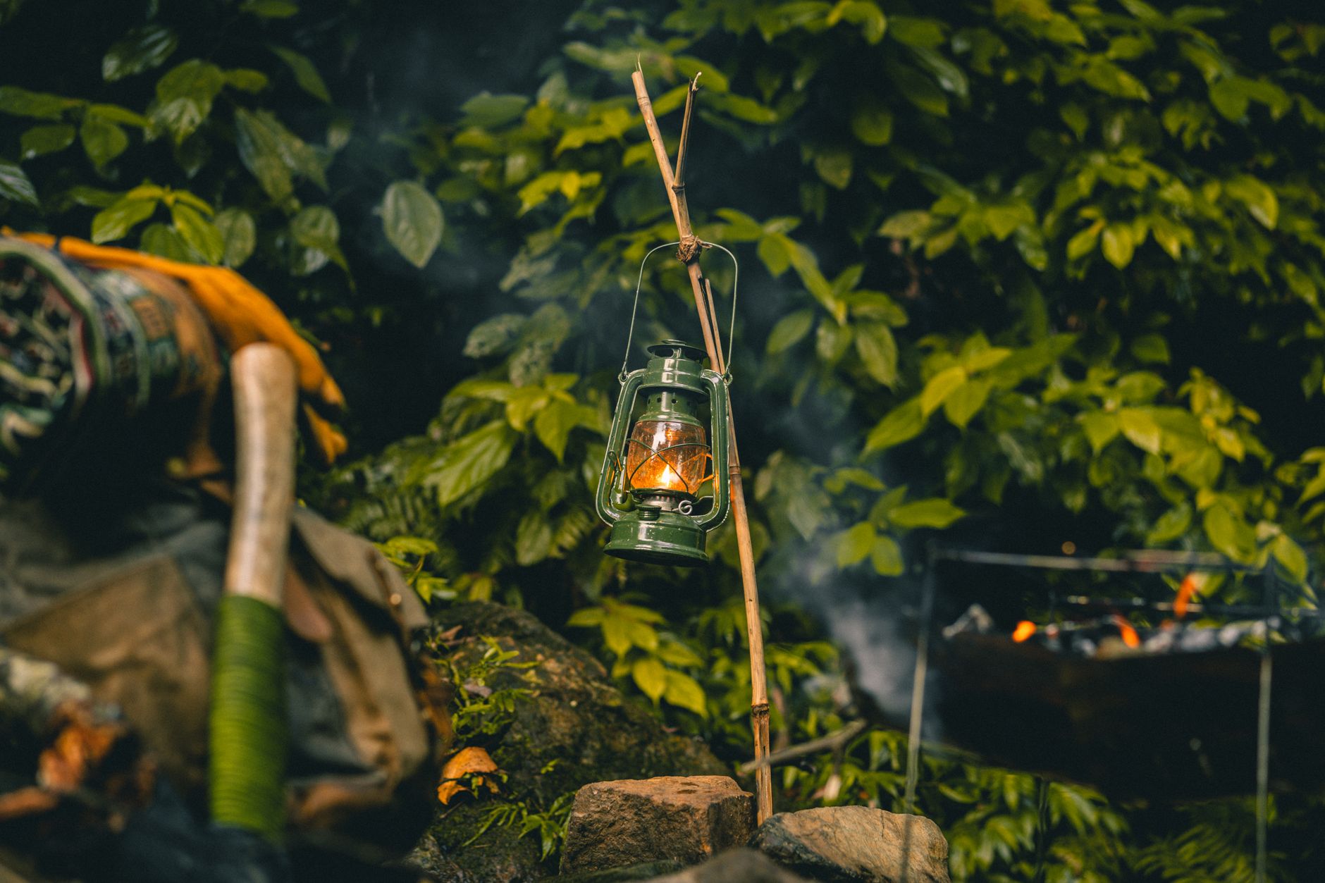 A rustic lantern hangs, casting warm light amidst lush foliage and camping gear.