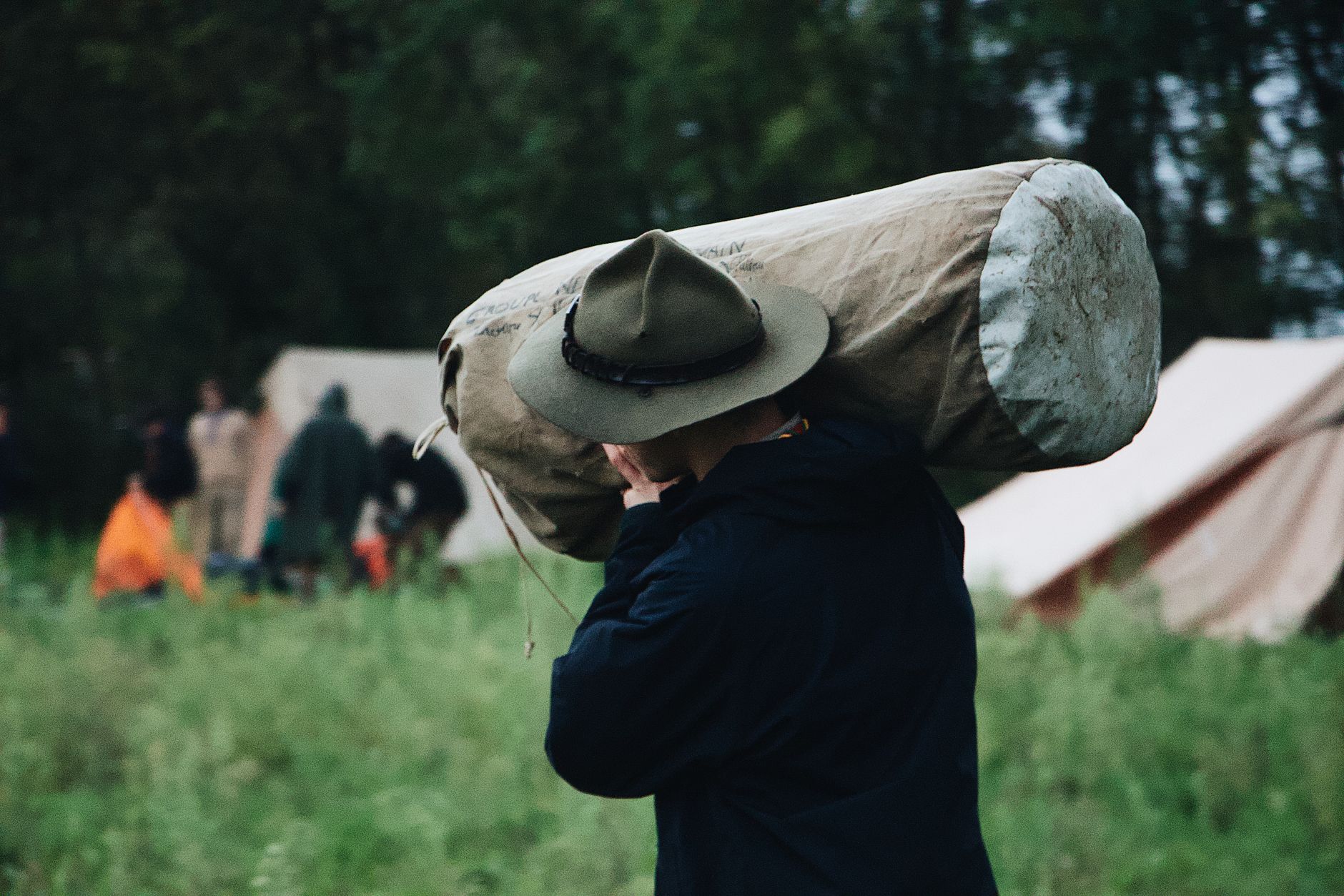 A man in a hat carries a large camping roll in a grassy field with tents.