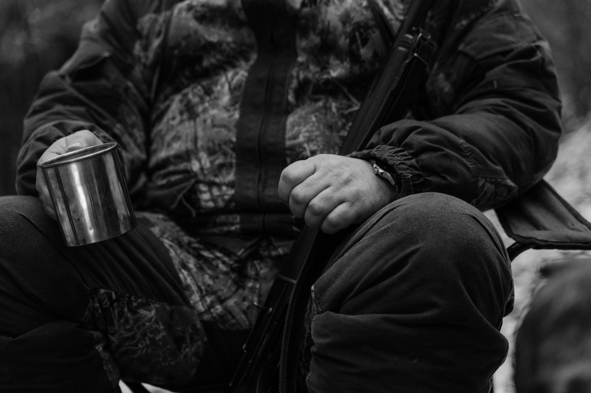 A hunter in camouflage sits holding a mug and rifle in a forest setting.