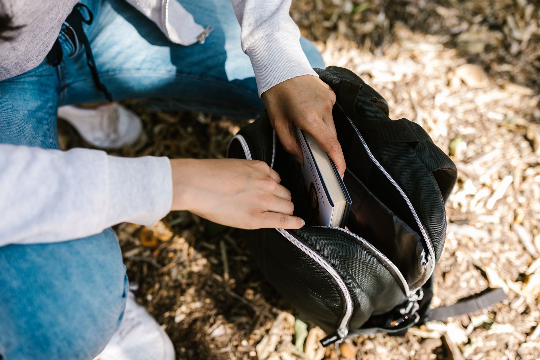 Close-up of hands placing a book in a backpack while seated outdoors.