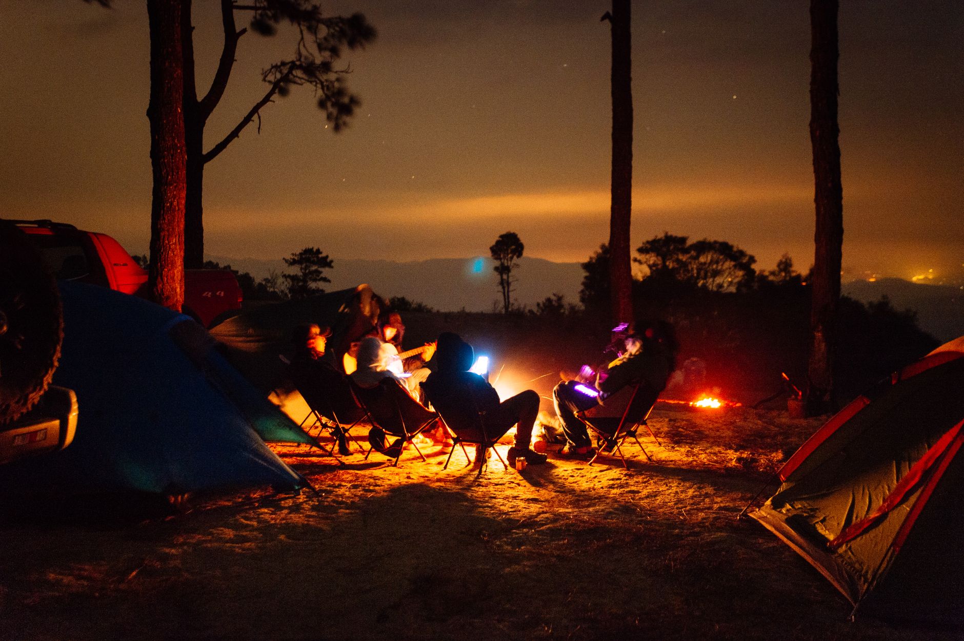 A group of adults enjoy a cozy campfire at a forest campsite under the night sky with tents and trees.