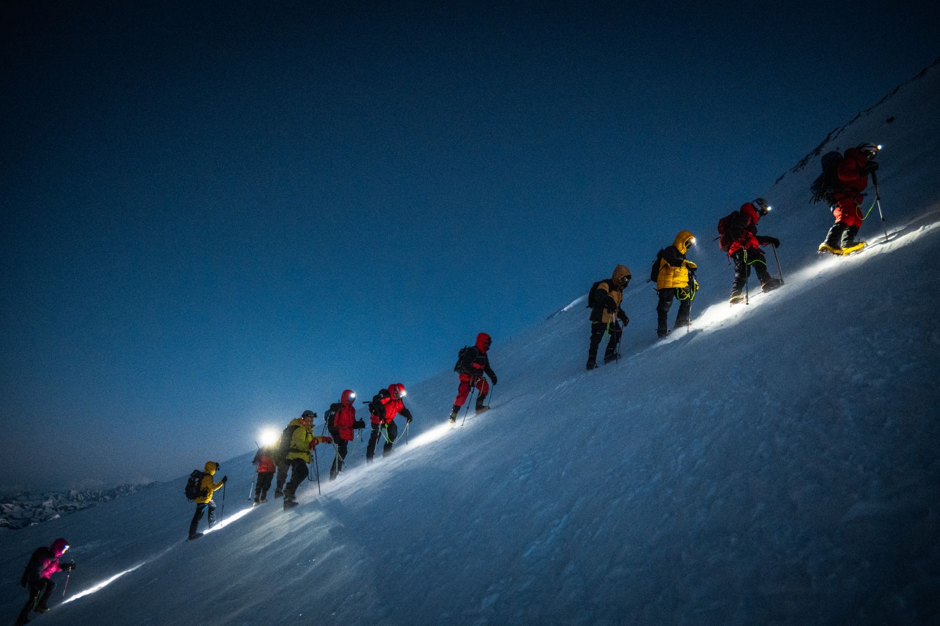 A group of climbers ascend Mount Elbrus at night, guided by headlamps, on a steep snowy slope.