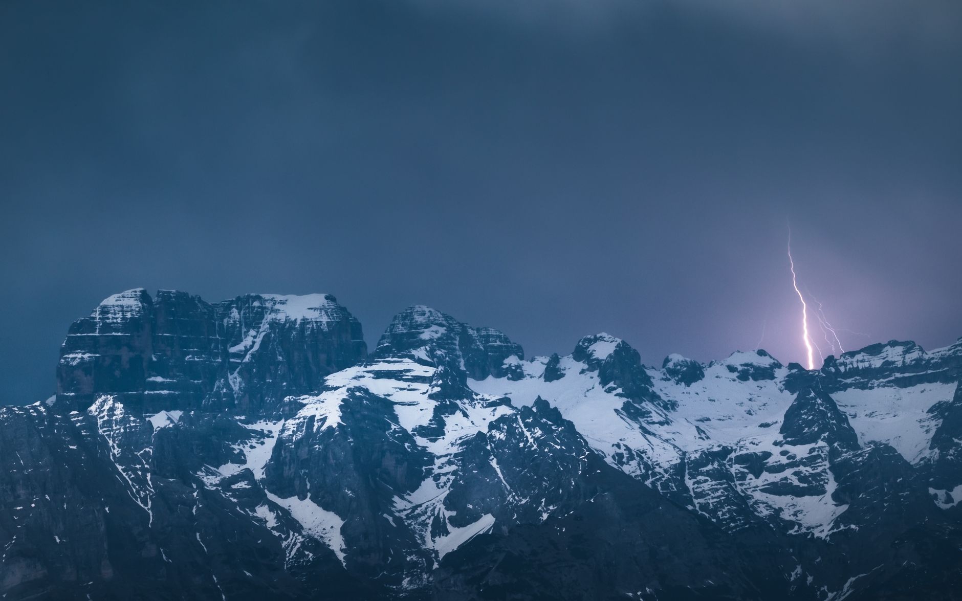 Lightning strikes over the snowy peaks of the Dolomites at night, creating a dramatic scene.
