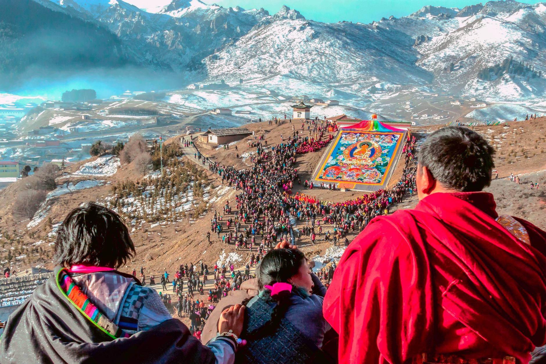 A vibrant Tibetan festival with monks and a large crowd in a snowy mountain setting.