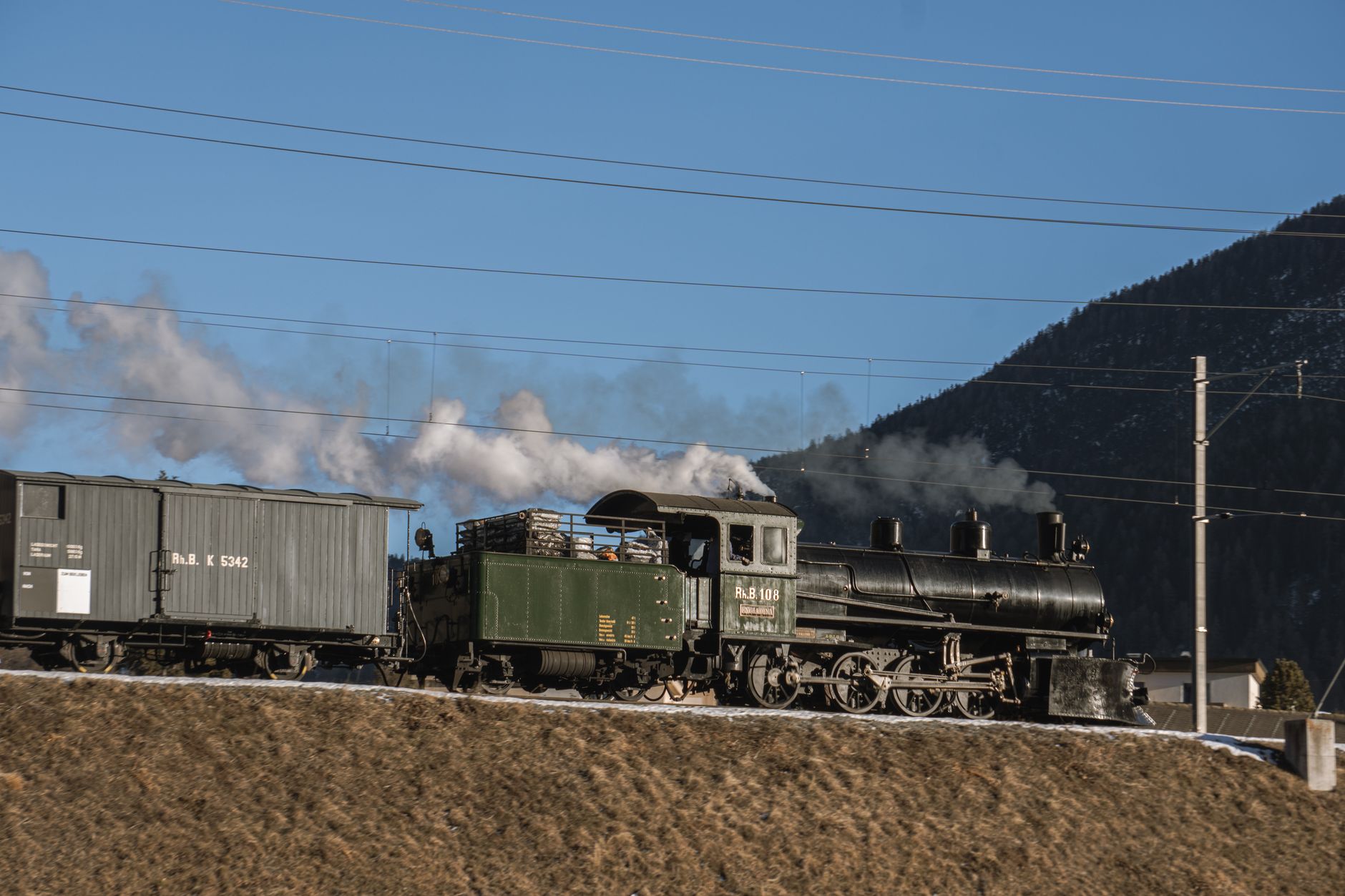 A vintage steam locomotive travels through a mountain landscape under a clear blue sky.
