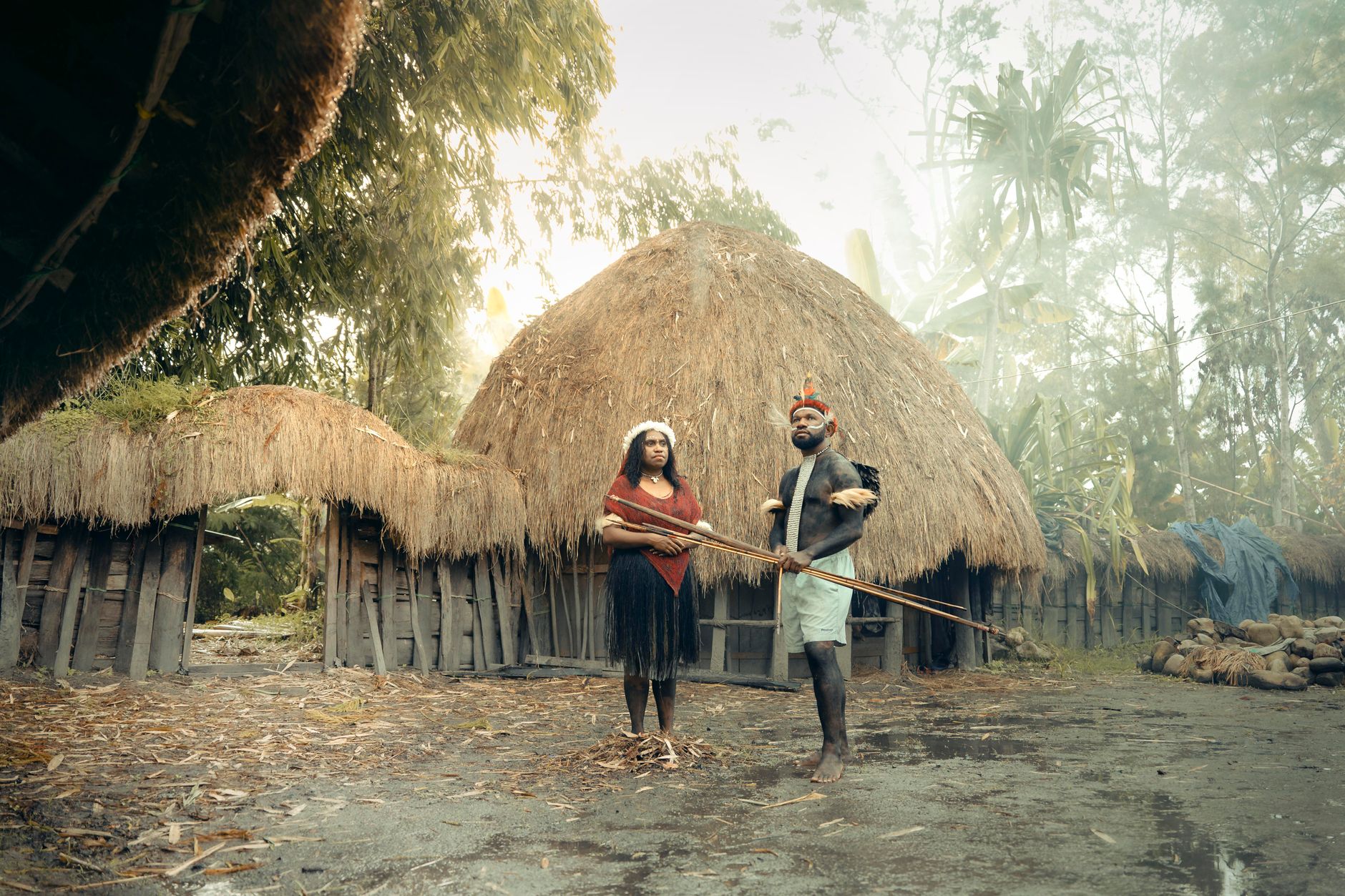 Two indigenous people in traditional attire stand in front of a thatched hut in Papua New Guinea.