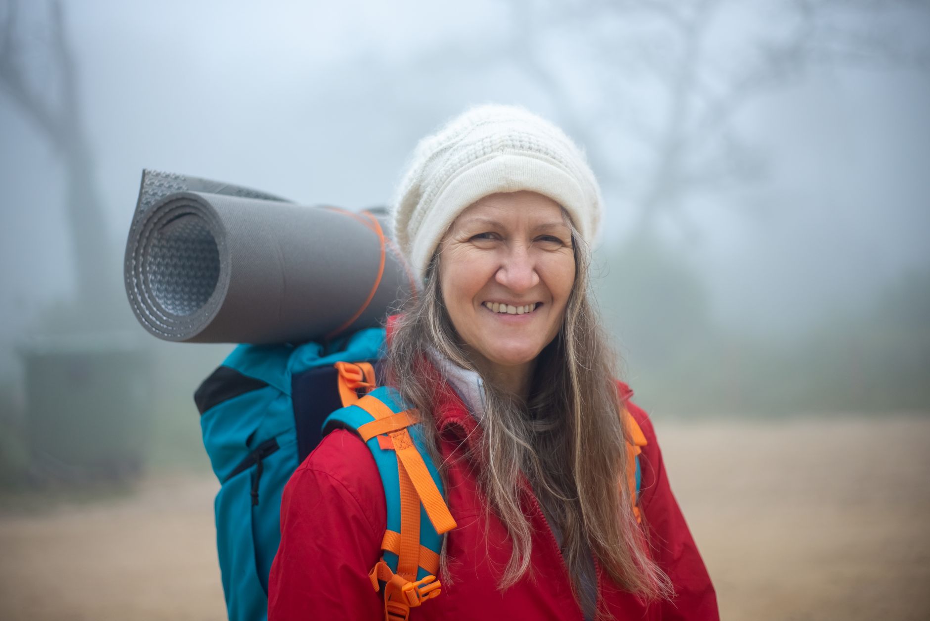 Elderly woman smiling while hiking in misty Portugal with backpack and winter gear.