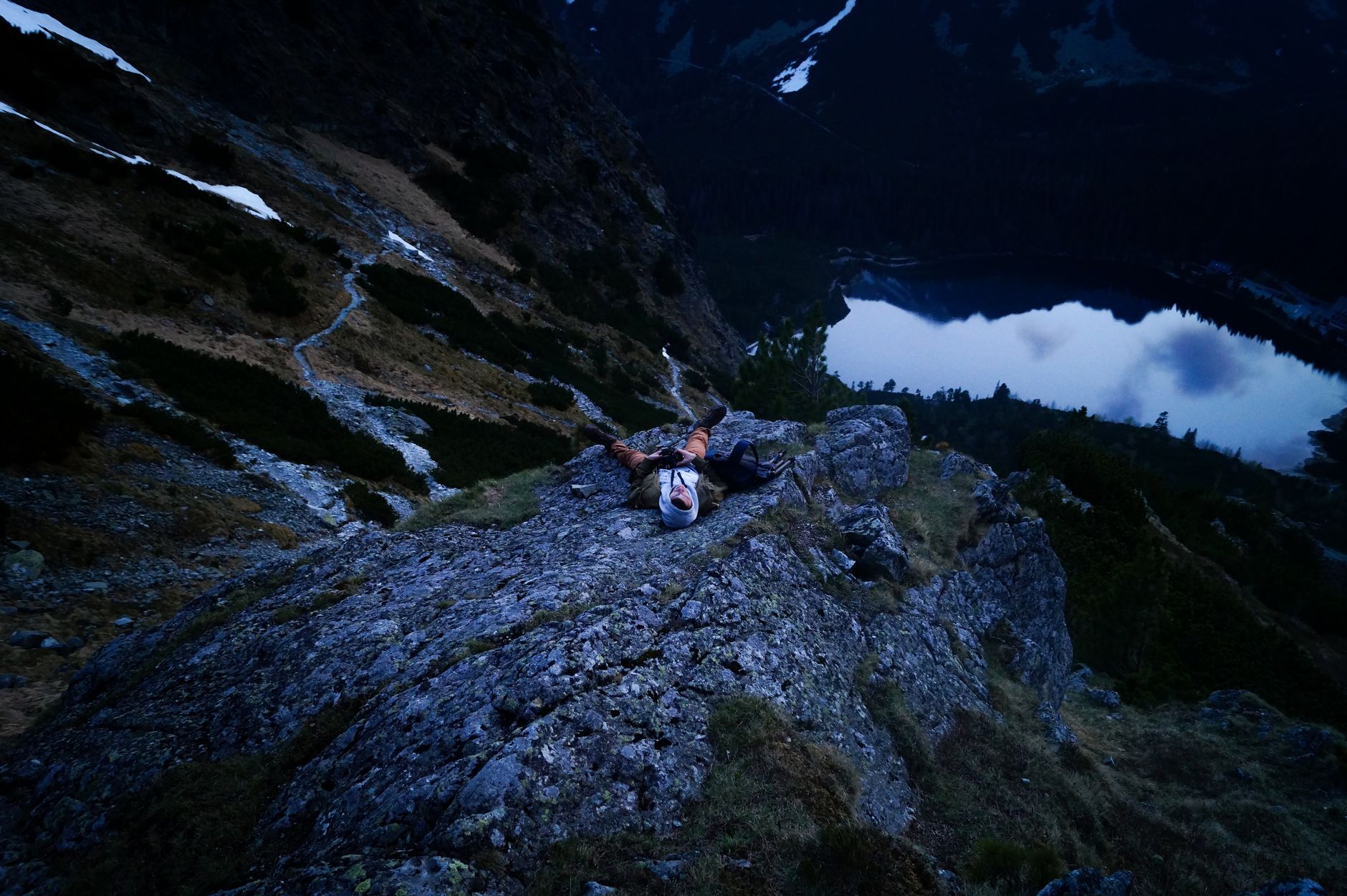 A lone hiker lies on a rock, enjoying the view of a tranquil lake in the mountains at twilight.