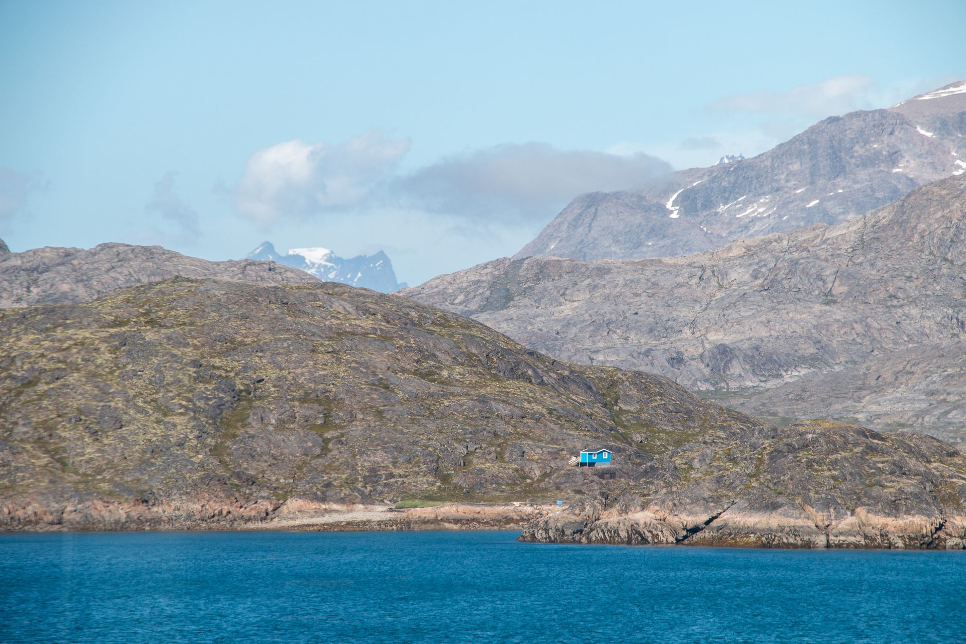 A solitary blue house set against Greenland's rugged mountains and clear waters, embodying isolation and natural beauty.
