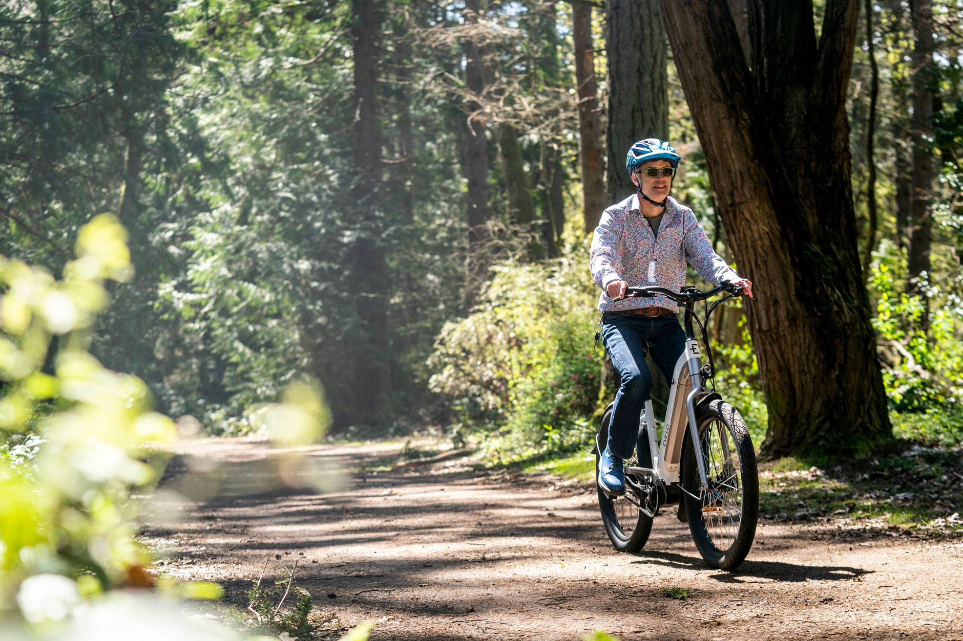 A man riding an e-bike through a forest path in Seattle, wearing a helmet and sunglasses.