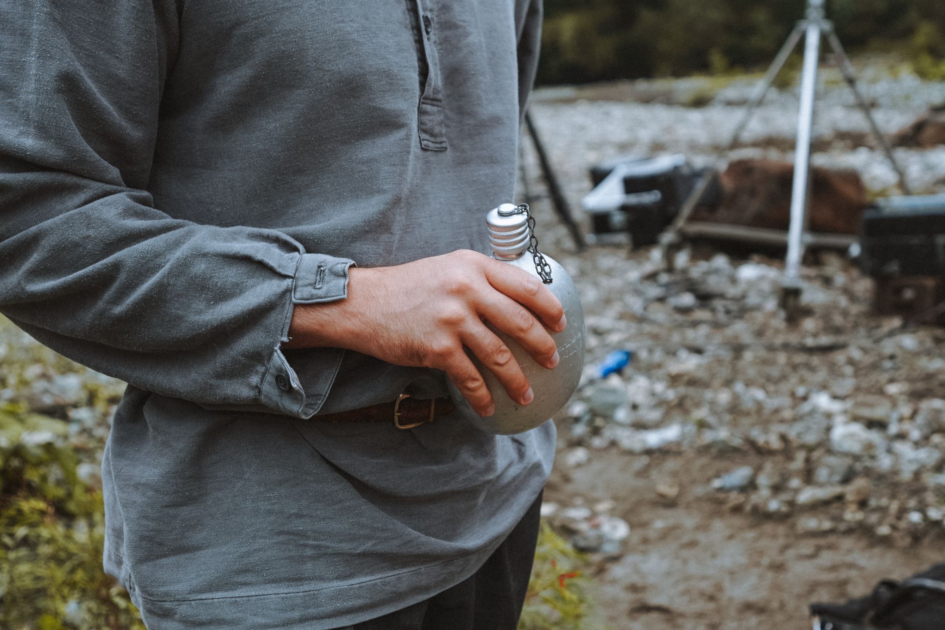 Casual scene of a man holding a tin bottle at a rocky outdoor campsite. Adventure vibe.
