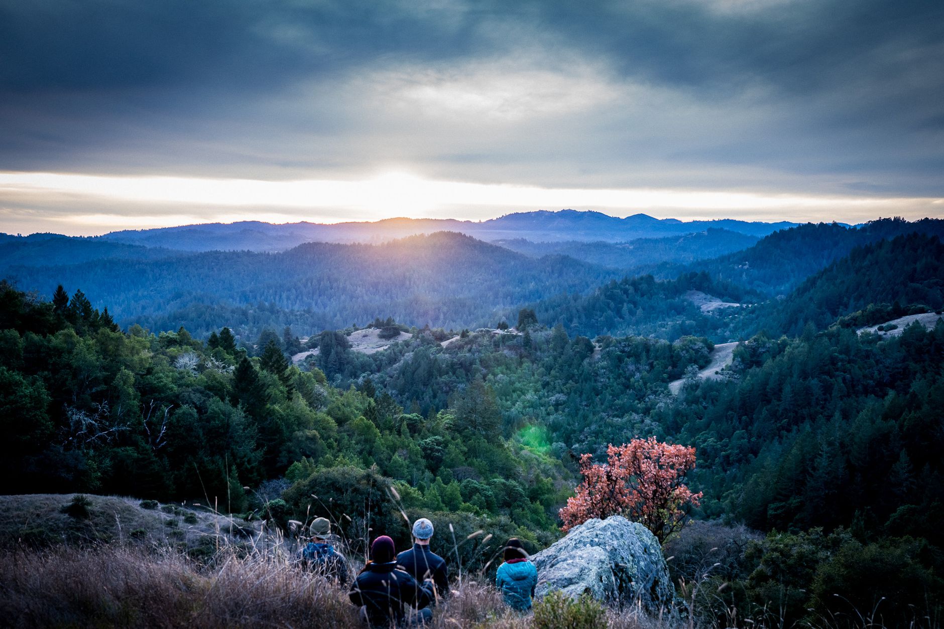 A group of hikers watches the sunrise over lush green mountains under a dramatic sky.