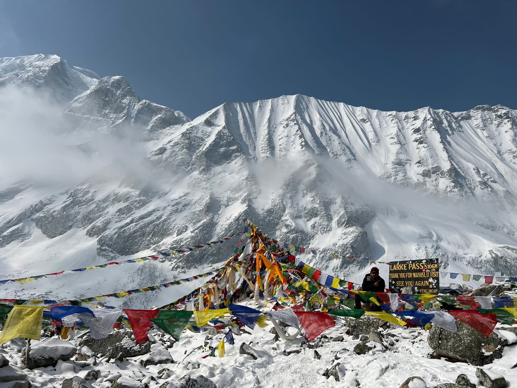 Colorful prayer flags flutter at snow-covered Larke Pass under majestic mountains.