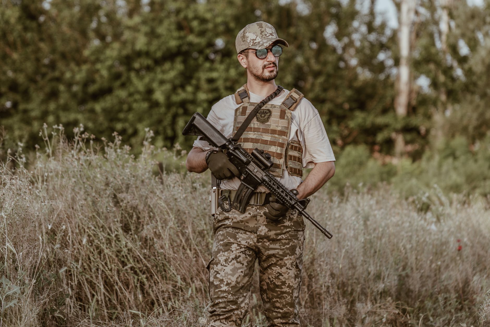 A soldier in camouflage uniform stands with a rifle in a rural grassland area.