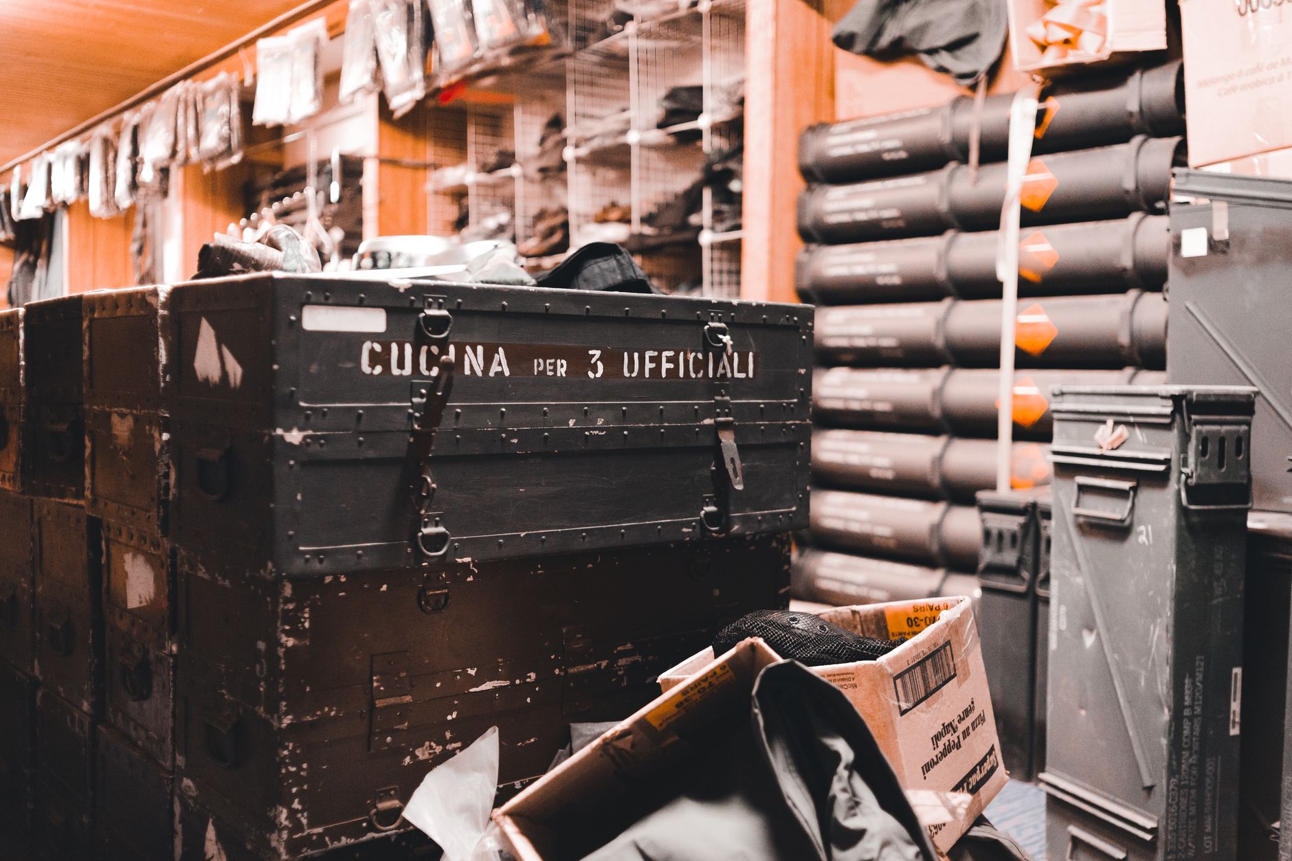 A busy military storage room filled with equipment and crates, labeled 'Cucina per 3 Ufficiali'.