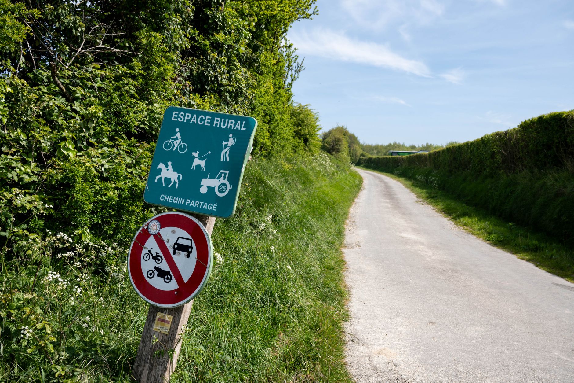 Rural road in Escalles with shared path sign, surrounded by lush greenery under clear skies.